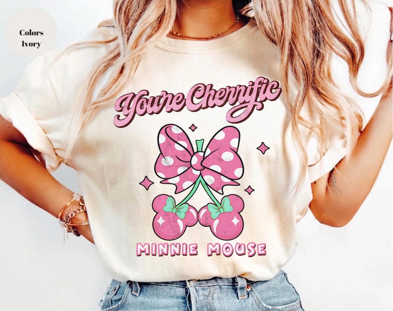 Pode incluir: Camiseta cor marfim com la&ccedil;o rosa de bolinhas brancas e design de cerejas. O texto "You're Cherrific" est&aacute; acima do la&ccedil;o e "Minnie Mouse" abaixo. A camiseta tem um corte folgado.