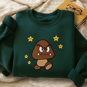 Puede incluir: Una sudadera verde oscuro con un personaje de dibujos animados Goomba bordado y acentos de estrellas amarillas. La sudadera tiene mangas largas y cuello redondo. La prenda se exhibe sobre una manta de color crema. El Goomba tiene una expresión enfadada.