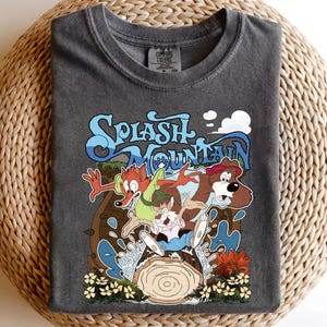 May include: Dark gray t-shirt featuring a colorful graphic of cartoon characters on a log flume ride. The design includes the words "Splash Mountain" in a stylized font, with images of a fox, bear, and rabbit. The shirt is made of a soft material.