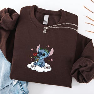 Puede incluir: Una sudadera marr&oacute;n con un personaje de dibujos animados bordado sosteniendo una rosa, sentado en una nube con corazones y sobres. Un collar de plata con un colgante est&aacute; drapeado sobre la sudadera. La sudadera est&aacute; doblada sobre unos vaqueros azules.