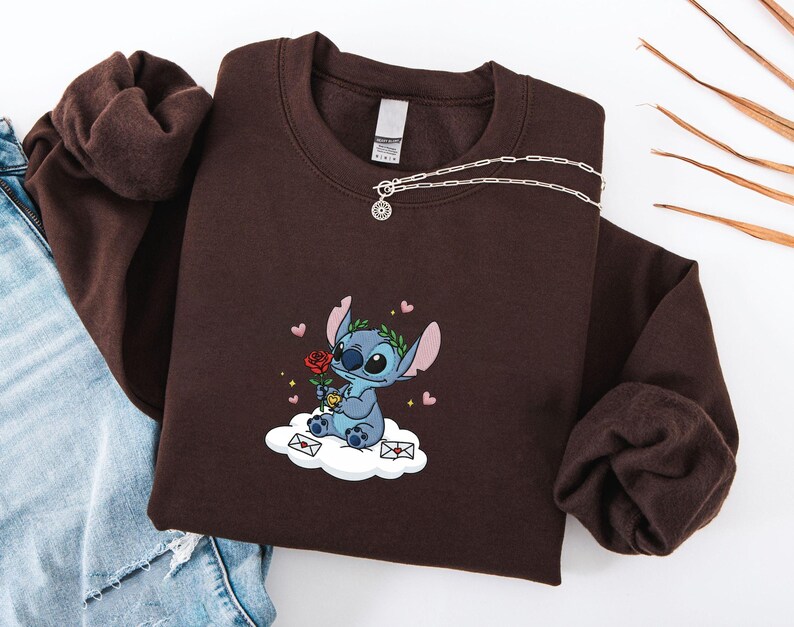 Puede incluir: Una sudadera marr&oacute;n con un personaje de dibujos animados bordado sosteniendo una rosa, sentado en una nube con corazones y sobres. Un collar de plata con un colgante est&aacute; drapeado sobre la sudadera. La sudadera est&aacute; doblada sobre unos vaqueros azules.