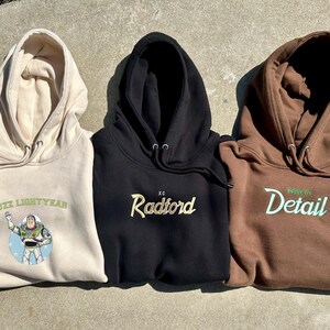 Puede incluir: Tres sudaderas con capucha en beige, negro y marr&oacute;n. La sudadera beige presenta un gr&aacute;fico de Buzz Lightyear. La sudadera negra tiene "Radford" bordado en dorado. La sudadera marr&oacute;n muestra "Drive By Detail" en verde.