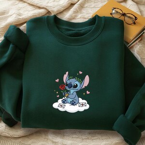 Puede incluir: Sudadera verde oscuro con una imagen bordada de un personaje de dibujos animados azul sosteniendo una rosa roja, sentado en una nube con corazones y sobres. El personaje tiene orejas grandes y una corona de laurel. Prenda c&oacute;moda.