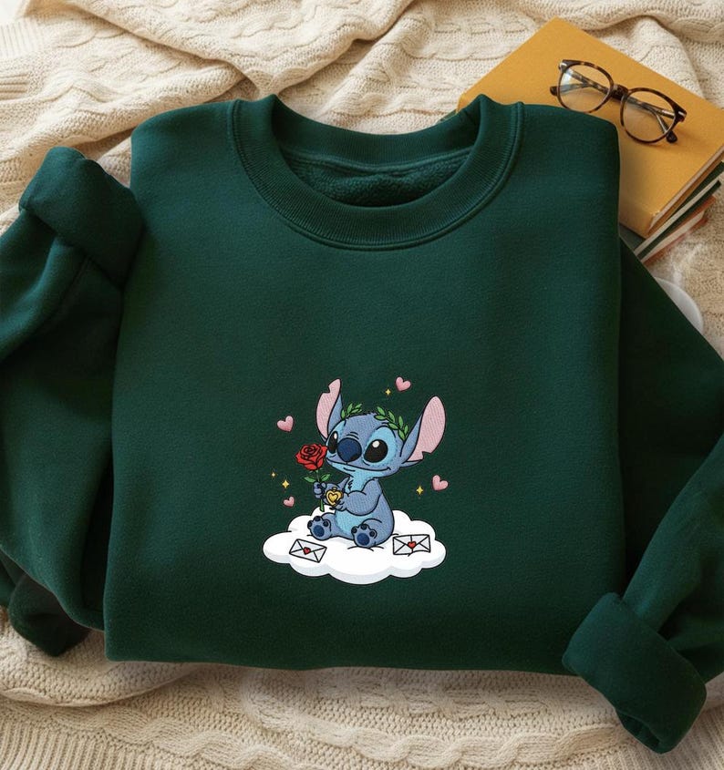 Puede incluir: Sudadera verde oscuro con una imagen bordada de un personaje de dibujos animados azul sosteniendo una rosa roja, sentado en una nube con corazones y sobres. El personaje tiene orejas grandes y una corona de laurel. Prenda c&oacute;moda.
