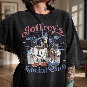 May include: A black short-sleeve t-shirt featuring a graphic print with the text Joffrey's since 1984 Social Club. The design includes a cartoon coffee cup and a coffee bean, both with mouse ears, standing in front of a castle illustration.