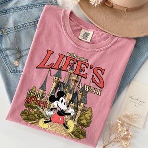Puede incluir: Camiseta rosa con un dibujo de Mickey Mouse corriendo frente a un castillo, con el texto "Life's a Walk in the Park" y "Walt Disney World". La camiseta est&aacute; doblada sobre unos vaqueros azules, con un collar dorado y un sombrero beige.