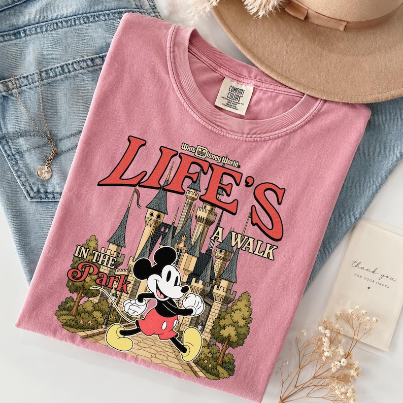 Puede incluir: Camiseta rosa con un dibujo de Mickey Mouse corriendo frente a un castillo, con el texto "Life's a Walk in the Park" y "Walt Disney World". La camiseta est&aacute; doblada sobre unos vaqueros azules, con un collar dorado y un sombrero beige.