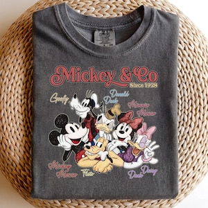 May include: Dark gray t-shirt featuring the text "Mickey & Co Since 1928" with a graphic of Mickey Mouse, Minnie Mouse, Donald Duck, Daisy Duck, Goofy, and Pluto. The shirt is made of a soft material.