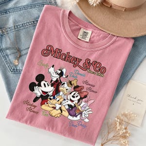 May include: A dusty rose-colored t-shirt featuring a graphic of Mickey Mouse and friends. The text "Mickey & Co Since 1928" is displayed above the characters. The shirt is folded on top of blue jeans.
