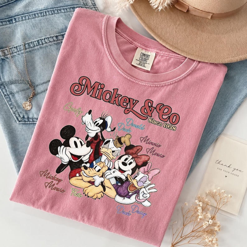 May include: A dusty rose-colored t-shirt featuring a graphic of Mickey Mouse and friends. The text "Mickey & Co Since 1928" is displayed above the characters. The shirt is folded on top of blue jeans.