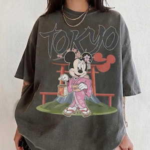 Peut inclure: T-shirt gris fonc&eacute; avec un motif de Minnie Mouse en kimono rose, tenant une lanterne, avec le mot "TOKYO" au-dessus. Le motif comprend un torii japonais et le mont Fuji.