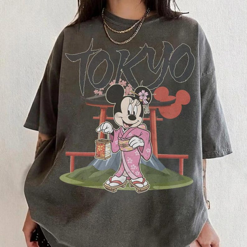 Peut inclure: T-shirt gris fonc&eacute; avec un motif de Minnie Mouse en kimono rose, tenant une lanterne, avec le mot "TOKYO" au-dessus. Le motif comprend un torii japonais et le mont Fuji.