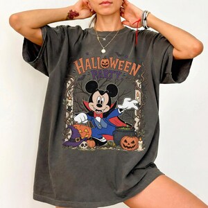 May include: A charcoal gray short-sleeve t-shirt featuring a graphic print of Mickey Mouse dressed as a vampire. The text HALLOWEEN PARTY is printed above the character, surrounded by pumpkins, a witch hat, a skull, and a cauldron in a forest.