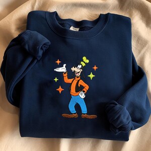 Puede incluir: Sudadera azul marino con un dise&ntilde;o bordado de Goofy. El personaje de dibujos animados lleva una camiseta naranja, pantalones azules y un sombrero verde. El dise&ntilde;o se complementa con estrellas de colores.