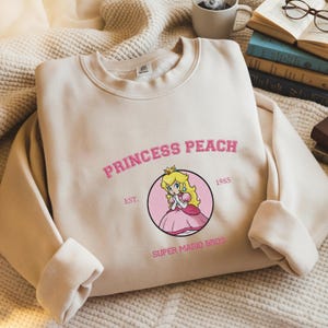 Peut inclure: Un sweat-shirt beige avec l'inscription rose "PRINCESS PEACH" et une illustration de la Princesse Peach de Super Mario Bros. Le design comprend également le texte "EST. 1985" et "SUPER MARIO BROS" en rose.