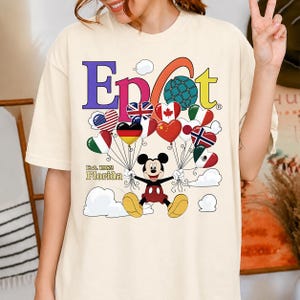 K&ouml;nnte beinhalten: Cremefarbenes T-Shirt mit dem Wort "Epcot" in bunten Buchstaben, einem Globus und herzf&ouml;rmigen Luftballons. Micky Maus h&auml;lt die Ballons, jeder mit einem Flaggen-Design. Der Text "Est. 1982 Florida" steht darunter.