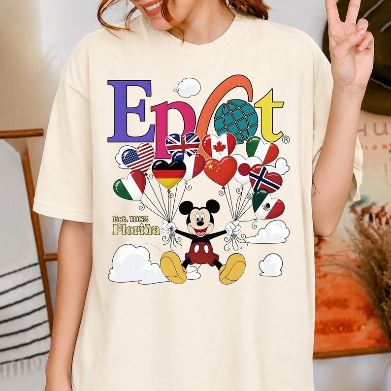 K&ouml;nnte beinhalten: Cremefarbenes T-Shirt mit dem Wort "Epcot" in bunten Buchstaben, einem Globus und herzf&ouml;rmigen Luftballons. Micky Maus h&auml;lt die Ballons, jeder mit einem Flaggen-Design. Der Text "Est. 1982 Florida" steht darunter.