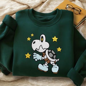 Puede incluir: Sudadera verde oscuro con un personaje de dibujos animados de tortuga esquelética bordada, caparazón gris, guantes y zapatos azul claro y estrellas amarillas. La sudadera está doblada sobre una manta color crema.