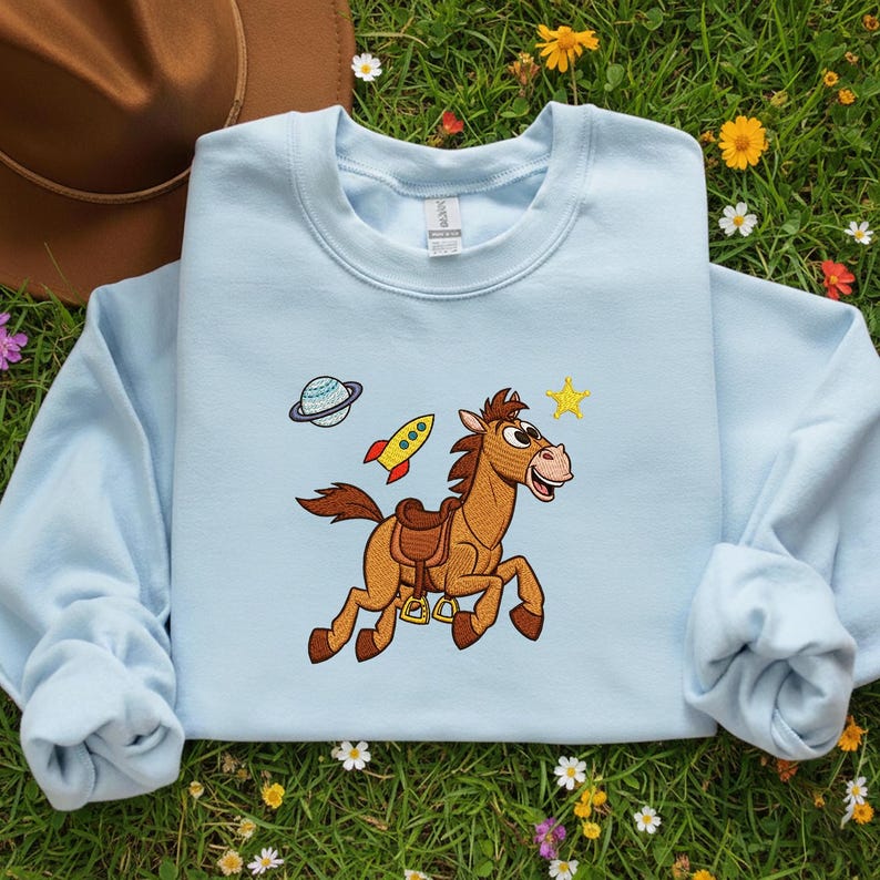 Pu&ograve; includere: Felpa azzurra con ricamo di un cavallo dei cartoni animati, un razzo, un pianeta e una stella. Il cavallo &egrave; marrone con una sella marrone e una bocca rosa. Il razzo &egrave; giallo e rosso. Il disegno &egrave; sul davanti.