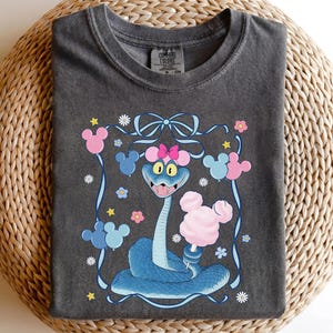 May include: Dark gray Comfort Colors t-shirt featuring a cartoon snake with a pink bow, holding cotton candy. The design includes Mickey Mouse-shaped balloons and flowers, with a blue ribbon border.