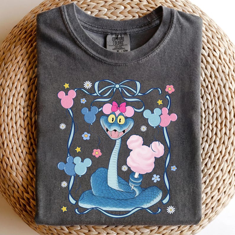 May include: Dark gray Comfort Colors t-shirt featuring a cartoon snake with a pink bow, holding cotton candy. The design includes Mickey Mouse-shaped balloons and flowers, with a blue ribbon border.