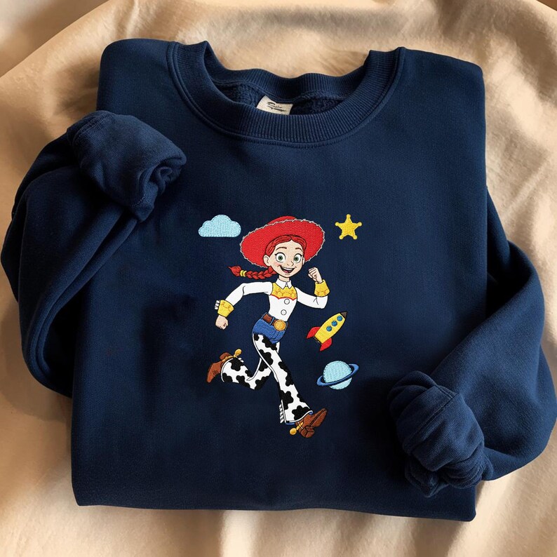 May include: Navy blue sweatshirt with an embroidered cartoon character in a red hat and cow-print trousers. The design includes a yellow star, a rocket, a planet, and a cloud. The sweatshirt has a crew neck and cuffed sleeves.