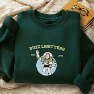 Puede incluir: Sudadera verde oscuro con un dise&ntilde;o de Buzz Lightyear de Toy Story. El dise&ntilde;o incluye el texto "Buzz Lightyear" sobre una ilustraci&oacute;n del personaje, con "EST. 1995" debajo. La sudadera est&aacute; doblada sobre una superficie color crema.