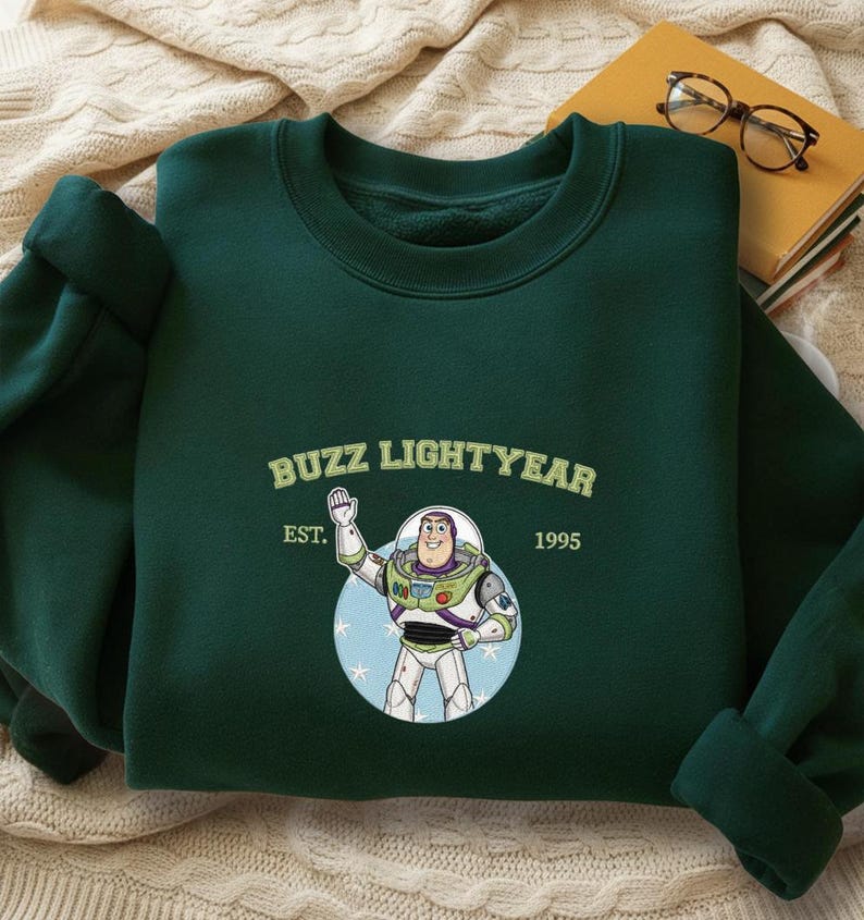 Puede incluir: Sudadera verde oscuro con un dise&ntilde;o de Buzz Lightyear de Toy Story. El dise&ntilde;o incluye el texto "Buzz Lightyear" sobre una ilustraci&oacute;n del personaje, con "EST. 1995" debajo. La sudadera est&aacute; doblada sobre una superficie color crema.