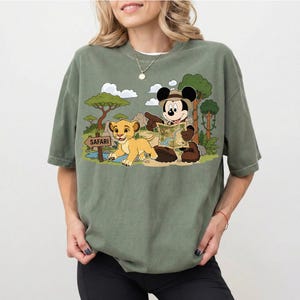 Op de afbeelding: Saliegroen T-shirt met een cartoonafbeelding van Mickey Mouse en Simba uit The Lion King op safari. Mickey Mouse houdt een kaart vast en Simba loopt in de buurt van een bord met "Safari".