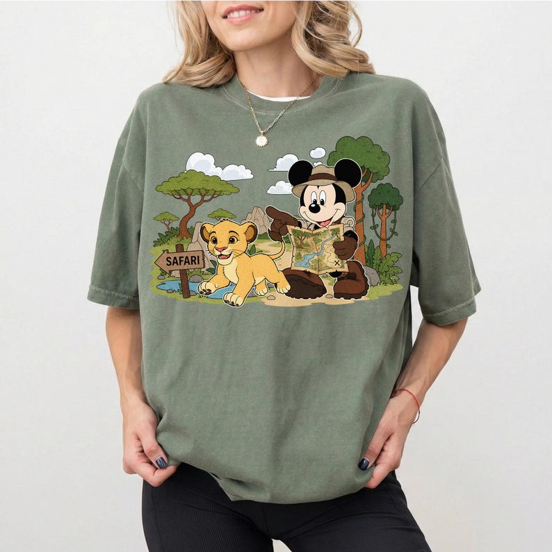 Op de afbeelding: Saliegroen T-shirt met een cartoonafbeelding van Mickey Mouse en Simba uit The Lion King op safari. Mickey Mouse houdt een kaart vast en Simba loopt in de buurt van een bord met "Safari".