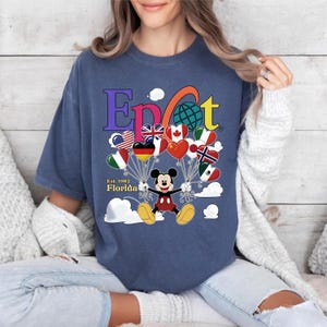 K&ouml;nnte beinhalten: Ein marineblaues T-Shirt mit dem Wort "Epcot" in bunten Buchstaben, einem Globus und Mickey Mouse, der herzf&ouml;rmige Ballons mit Flaggen h&auml;lt. Der Text "Est. 1982 Florida" ist ebenfalls sichtbar.