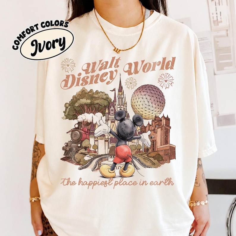 May include: Ivory-colored t-shirt with a vintage-style Walt Disney World graphic. The design features Mickey Mouse, a train, a castle, and the text "Walt Disney World" and "the happiest place on earth". The shirt has "Comfort Colors" printed on it.