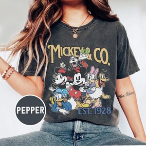 May include: Dark gray t-shirt featuring a graphic of Mickey Mouse and friends, with the text "Mickey & Co. EST. 1928" in yellow. The design includes Minnie Mouse, Donald Duck, Daisy Duck, Goofy, and Pluto.