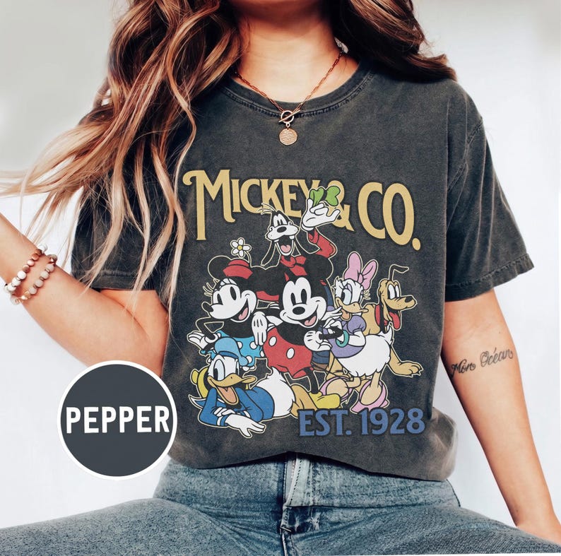May include: Dark gray t-shirt featuring a graphic of Mickey Mouse and friends, with the text "Mickey & Co. EST. 1928" in yellow. The design includes Minnie Mouse, Donald Duck, Daisy Duck, Goofy, and Pluto.