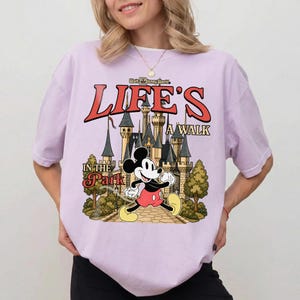 Puede incluir: Camiseta lila claro con Mickey Mouse frente a un castillo y el texto "Life's a Walk in the Park" en rojo y beige. La camiseta es de corte holgado.