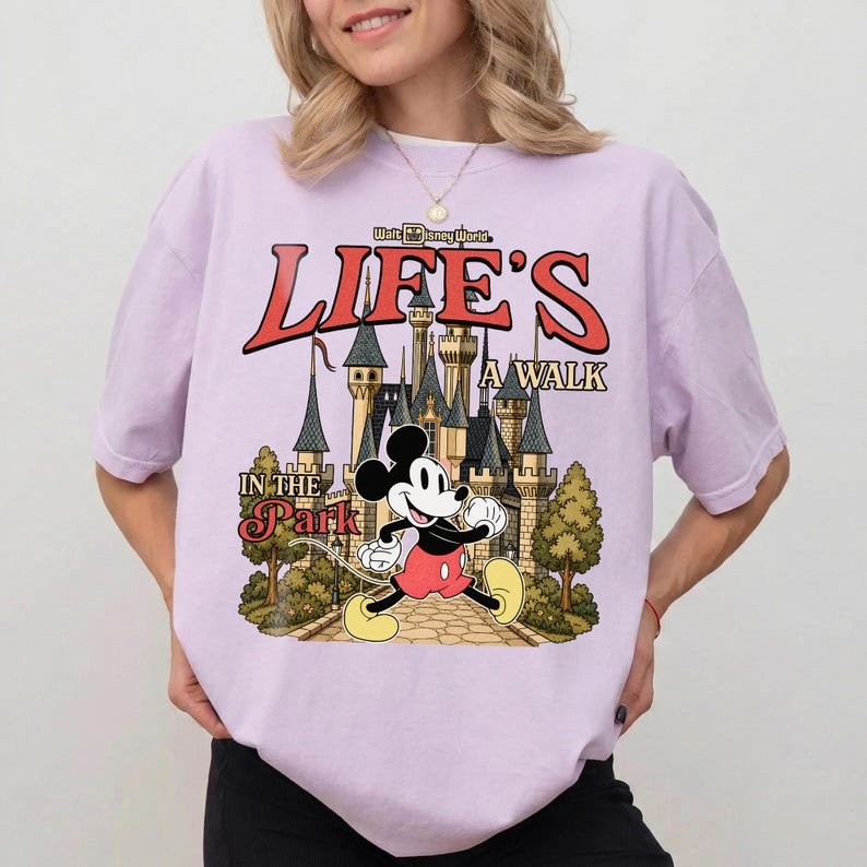Puede incluir: Camiseta lila claro con Mickey Mouse frente a un castillo y el texto "Life's a Walk in the Park" en rojo y beige. La camiseta es de corte holgado.