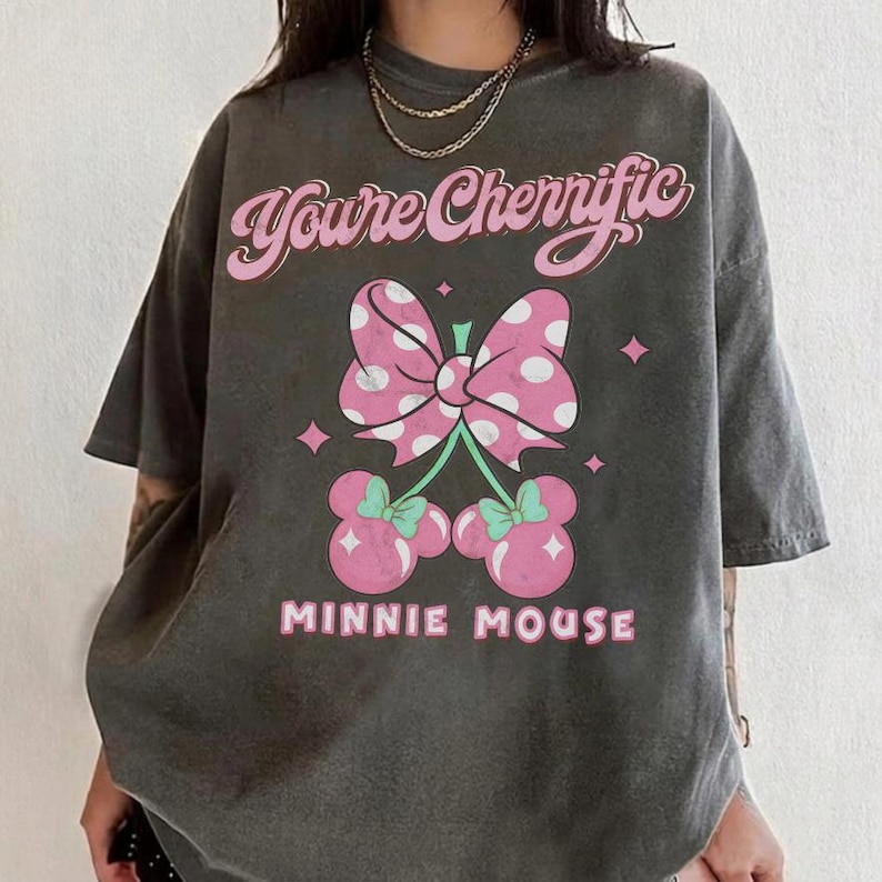 Pode incluir: T-shirt cinza escuro com la&ccedil;o rosa e branco de bolinhas e design de cereja da Minnie Mouse. O texto "You're Cherrific" e "MINNIE MOUSE" s&atilde;o em rosa. A camisa tem mangas curtas.