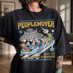 May include: Black t-shirt featuring a graphic of the Tomorrowland PeopleMover ride with Mickey, Minnie, Donald, Daisy, and Goofy. The shirt includes the text "TOMORROWLAND TRANSIT AUTHORITY PEOPLEMOVER" and "KEEP MOVING FORWARD TO A NEW TOMORROW."