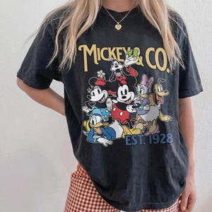 May include: Black t-shirt featuring a graphic of Mickey Mouse and friends, with the text "Mickey & Co. EST. 1928" in yellow and blue. The design includes Mickey, Minnie, Donald, Daisy, Goofy, and Pluto.
