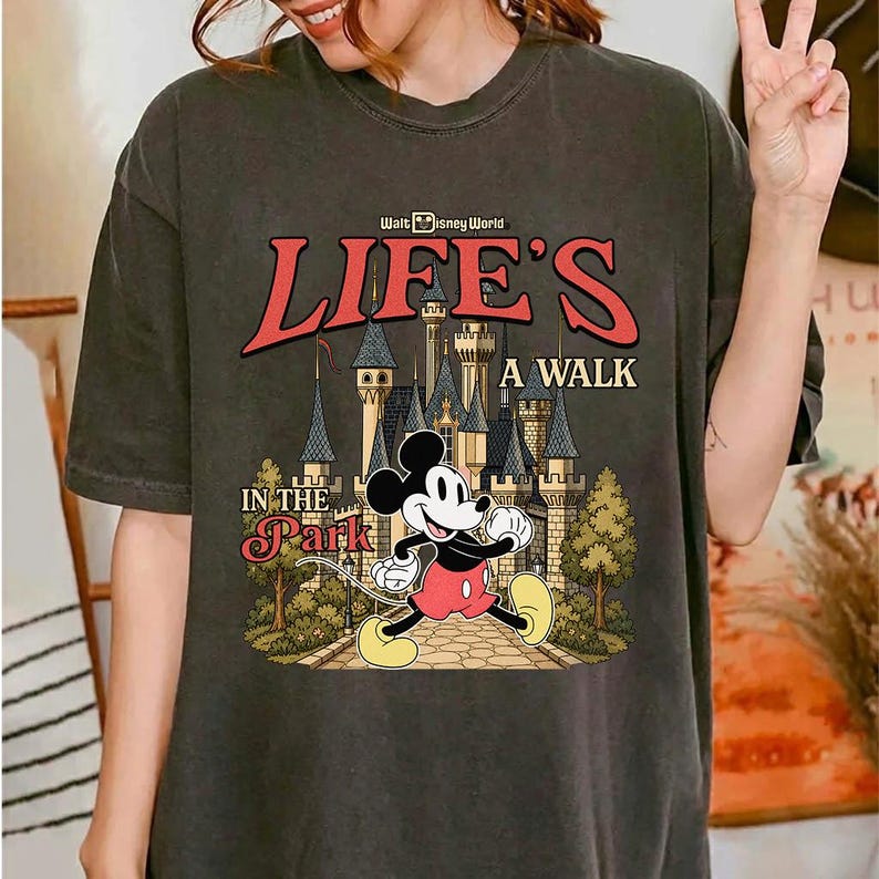 Puede incluir: Camiseta gris oscuro con Mickey Mouse caminando frente a un castillo. La camiseta tiene el texto "Walt Disney World" encima de las palabras "LIFE'S A WALK IN THE PARK" en rojo y blanco.