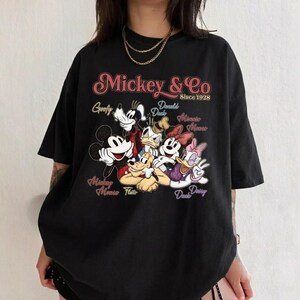 May include: Black t-shirt featuring a graphic of Mickey Mouse and friends. The design includes Mickey, Minnie, Donald, Daisy, Goofy, and Pluto, with the text "Mickey & Co Since 1928". The shirt has a vintage, retro style.