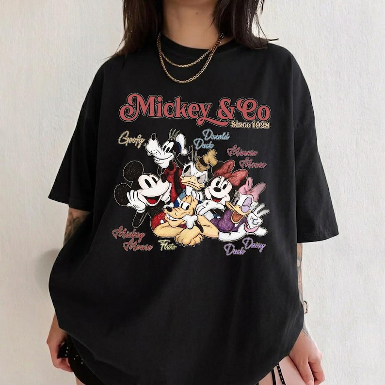 May include: Black t-shirt featuring a graphic of Mickey Mouse and friends. The design includes Mickey, Minnie, Donald, Daisy, Goofy, and Pluto, with the text "Mickey & Co Since 1928". The shirt has a vintage, retro style.