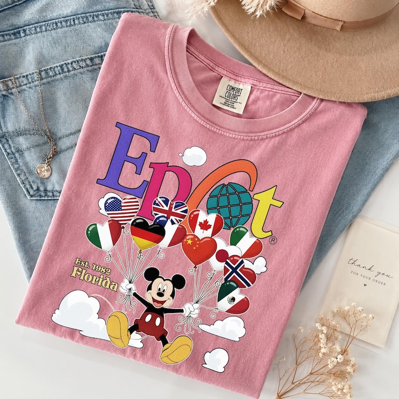 K&ouml;nnte beinhalten: Hellrosa T-Shirt mit farbenfroher Grafik. Das Design zeigt das Wort "EPCOT", Mickey Mouse, der herzf&ouml;rmige Ballons mit Flaggen h&auml;lt, und den Text "Est. 1982 Florida".
