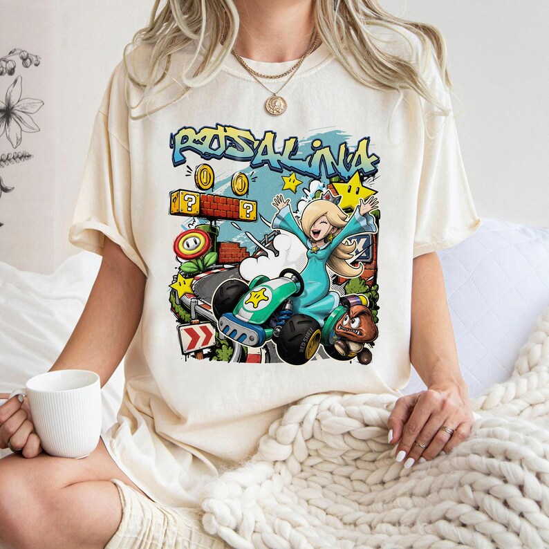 May include: Cream-colored t-shirt featuring a colorful graphic of Rosalina from Mario Kart. Rosalina is in a green kart, with the word "Rosalina" in graffiti-style lettering above her. The design includes stars, coins, and a Goomba.