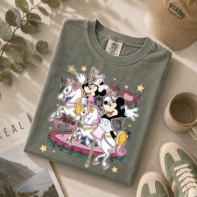Puede incluir: Una camiseta verde salvia con un dise&ntilde;o de carrusel con los personajes de Mickey y Minnie Mouse montados en caballos blancos. El dise&ntilde;o incluye detalles en rosa, amarillo y dorado. Tambi&eacute;n se ven una gorra beige, una taza y zapatillas.