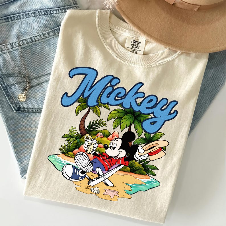May include: Cream-colored t-shirt featuring a graphic of Mickey Mouse relaxing on a beach with palm trees and the word "Mickey" in blue script. The shirt is folded on top of blue jeans and a tan hat.