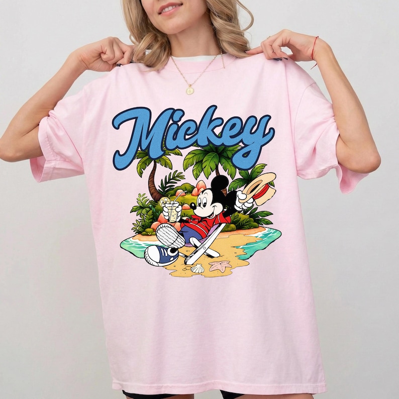 May include: Pink t-shirt featuring the word "Mickey" in blue script above a cartoon illustration of Mickey Mouse relaxing on a beach. Mickey is wearing a red shirt, blue shorts, and a straw hat, holding a drink.