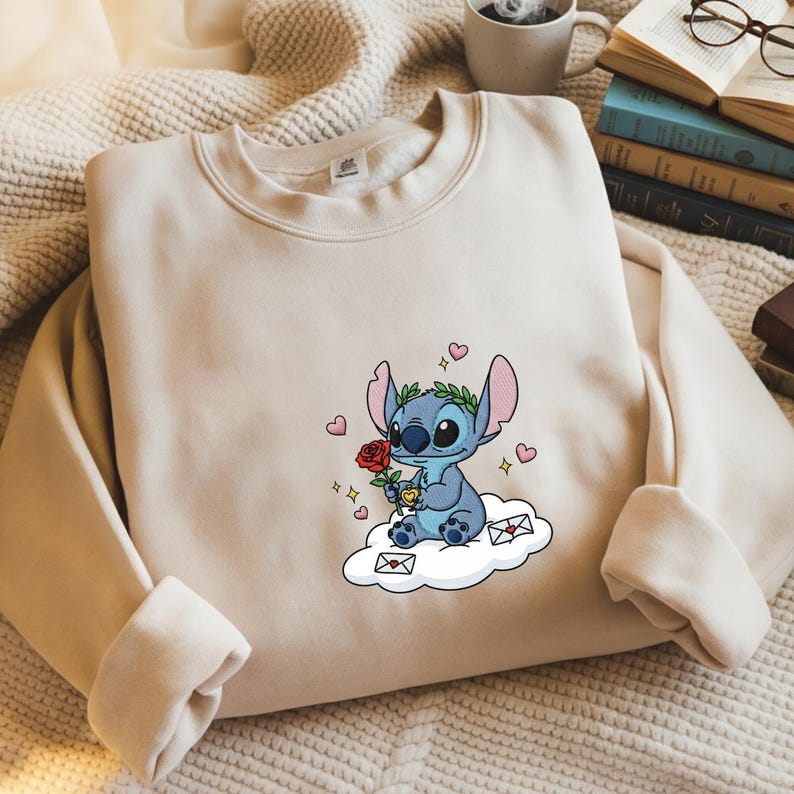 Puede incluir: Sudadera beige con un personaje de dibujos animados azul sosteniendo una rosa roja y un coraz&oacute;n. El personaje est&aacute; sentado en una nube con gr&aacute;ficos de coraz&oacute;n y sobre. La sudadera tiene cuello redondo y mangas largas.