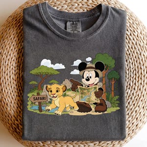 Op de afbeelding: Donkergrijs T-shirt met een cartoon safari sc&egrave;ne met Mickey Mouse, Simba en een kaart. Het ontwerp omvat bomen, een bord met de tekst "Safari" en wolken. Het shirt is gemaakt van zacht materiaal.