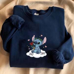Puede incluir: Sudadera azul marino con un personaje de dibujos animados bordado sosteniendo una rosa roja. El personaje azul est&aacute; sentado en una nube con corazones y sobres. Cuello redondo y mangas largas.
